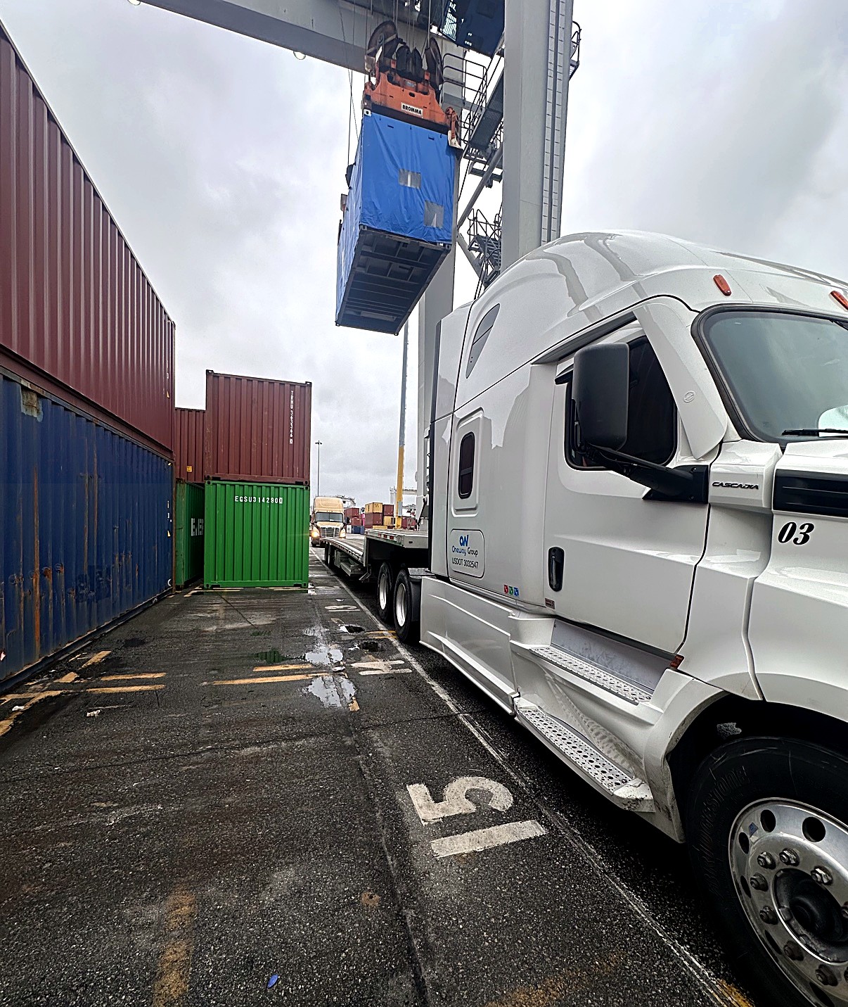 Oneway Group Cascadia unit 03 at a container port as a crane lifts a blue container over its flatbed, containers stacked alongside
